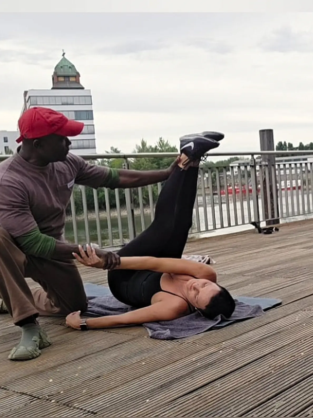 Trainer führt auf einem Holzsteg am Wasser eine Dehnübung mit einer Kundin